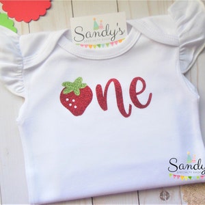 May include: White baby onesie with red glitter 'one' and a red glitter strawberry with green leaves.