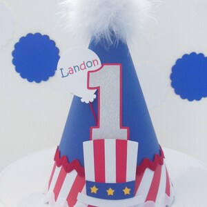 May include: A blue cone-shaped party hat topped with a white feather pom-pom. The hat features a large silver glitter number one, a red and white striped top hat graphic with three yellow stars, and a white balloon cutout with the name Landon.