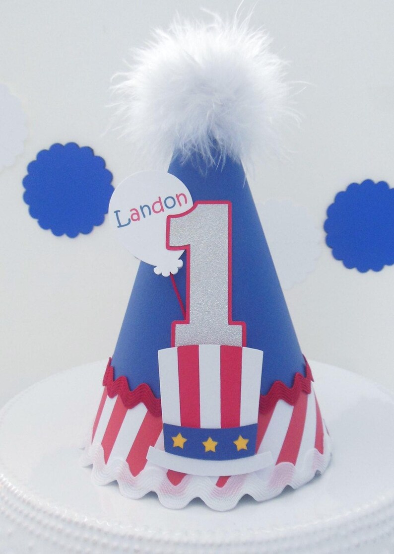 May include: A blue cone-shaped party hat topped with a white feather pom-pom. The hat features a large silver glitter number one, a red and white striped top hat graphic with three yellow stars, and a white balloon cutout with the name Landon.