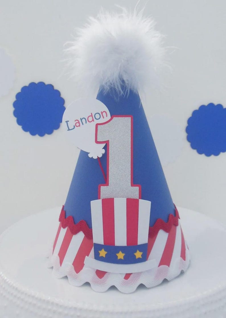 May include: A blue cone-shaped party hat topped with white feathers. It features a silver glitter number one, a red and white striped top hat with three yellow stars, and a white balloon cutout with the name Landon printed in blue text.