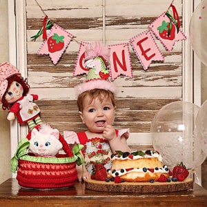 May include: A baby girl wearing a pink and green strawberry-themed dress and a pink and white party hat sits at a table with a strawberry shortcake cake, a basket with a stuffed white cat, and a doll. A banner with the word "ONE" is hanging above the table.
