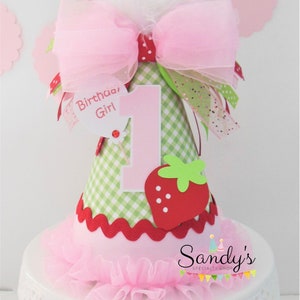 May include: A pink and green birthday hat with a strawberry design and a pink bow. The hat says "Birthday Girl" and has the number "1" on it.