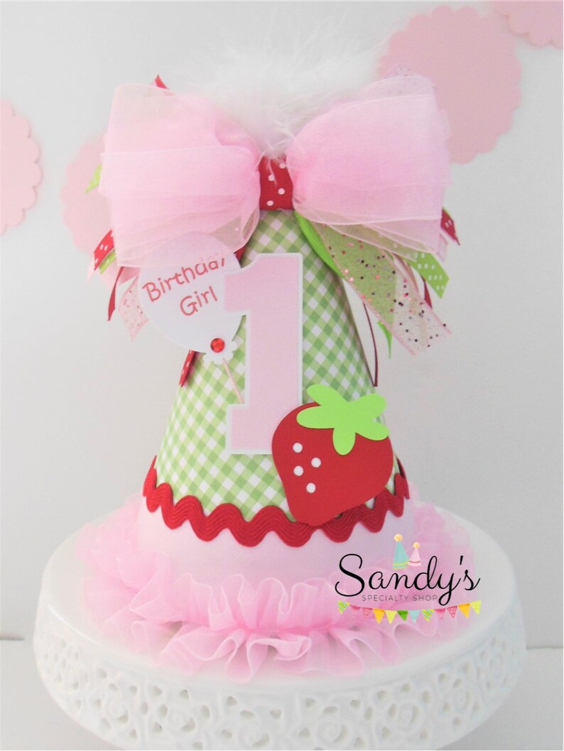 May include: A pink and green birthday hat with a strawberry design and a pink bow. The hat says "Birthday Girl" and has the number "1" on it.