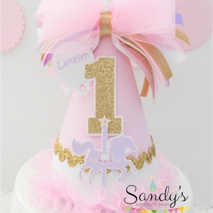 May include: Pink and gold birthday party hat with a white feather, a large pink bow, and a gold glitter number one. The hat has a purple and white carousel horse design and a white tag with the name "Lakelyn" written in purple.