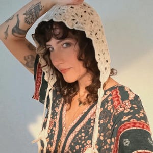 以下が含まれることがあります： クリーム色の、スカラップエッジと長い紐が付いたかぎ針編みのフード。 フードは、花柄と赤いアクセントが付いた濃いティール色のドレスの上に着用されています。 ドレスはVネックで長袖です。