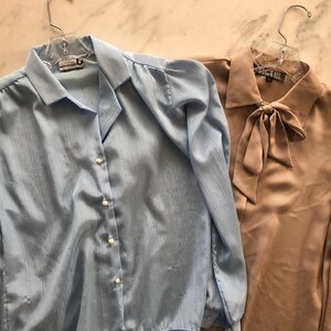 May include: Two long-sleeved blouses, one light blue with a button-up front and the other a beige color with a bow tie at the neck.
