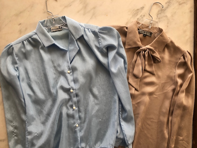May include: Two long-sleeved blouses, one light blue with a button-up front and the other a beige color with a bow tie at the neck.