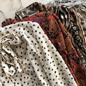 May include: A collection of five vintage blouses with different patterns. The blouses are hanging on hangers and are made of a variety of fabrics. The patterns include a white blouse with black polka dots, a red and black paisley print, a black and white zebra print, a brown and white floral print, and a beige and brown floral print.