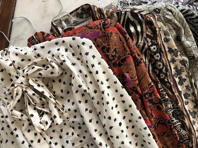 May include: A collection of five vintage blouses with different patterns. The blouses are hanging on hangers and are made of a variety of fabrics. The patterns include a white blouse with black polka dots, a red and black paisley print, a black and white zebra print, a brown and white floral print, and a beige and brown floral print.