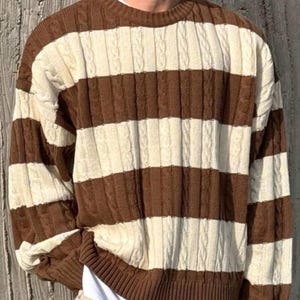 May include: A brown and cream-colored striped sweater with a cable knit design. The sweater has a crew neck and long sleeves. The stripes are horizontal and evenly spaced, creating a classic, cozy look.