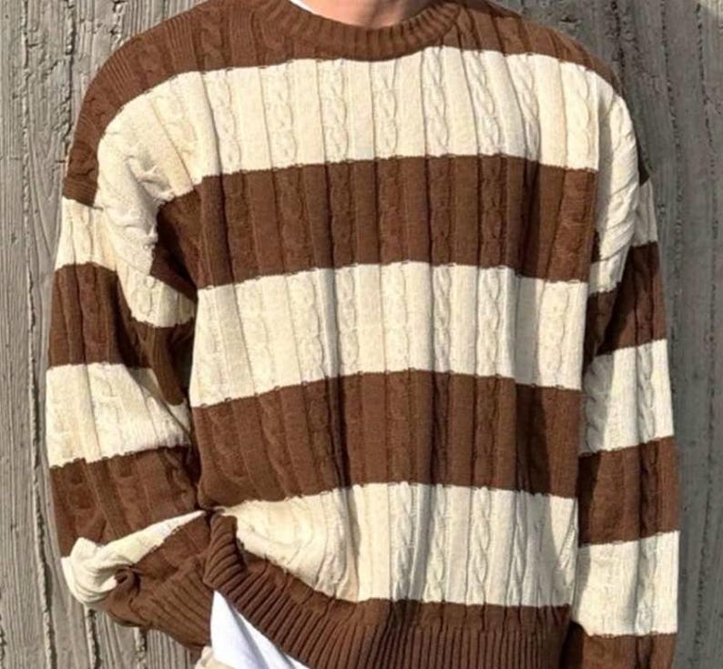 May include: A brown and cream-colored striped sweater with a cable knit design. The sweater has a crew neck and long sleeves. The stripes are horizontal and evenly spaced, creating a classic, cozy look.