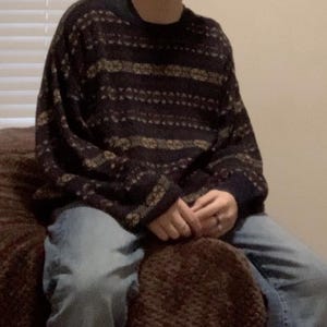 May include: A dark navy blue sweater with a repeating pattern of gold and tan horizontal stripes. The sweater has a crew neck and long sleeves. The person is wearing light blue jeans.
