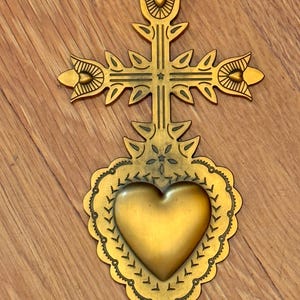 May include: A gold-colored metal cross with a heart-shaped emblem. The cross features ornate floral and leaf designs. The heart is centered below the cross, surrounded by a scalloped border. The piece is displayed on a wooden surface.