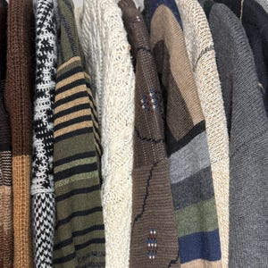 May include: A collection of various sweaters hanging on a rack. The sweaters display a range of colors and patterns, including stripes, cable knits, and geometric designs. The color palette includes brown, beige, green, and gray tones, offering a variety of textures and styles.