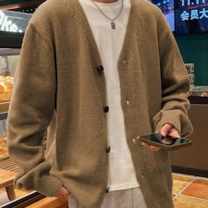 May include: A brown cardigan sweater with a V-neck and button closure. The sweater is worn over a white t-shirt. The person is holding a black smartphone. The background includes a bakery display and a sign with Chinese characters.