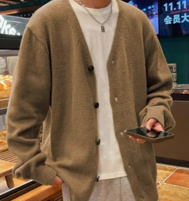 May include: A brown cardigan sweater with a V-neck and button closure. The sweater is worn over a white t-shirt. The person is holding a black smartphone. The background includes a bakery display and a sign with Chinese characters.