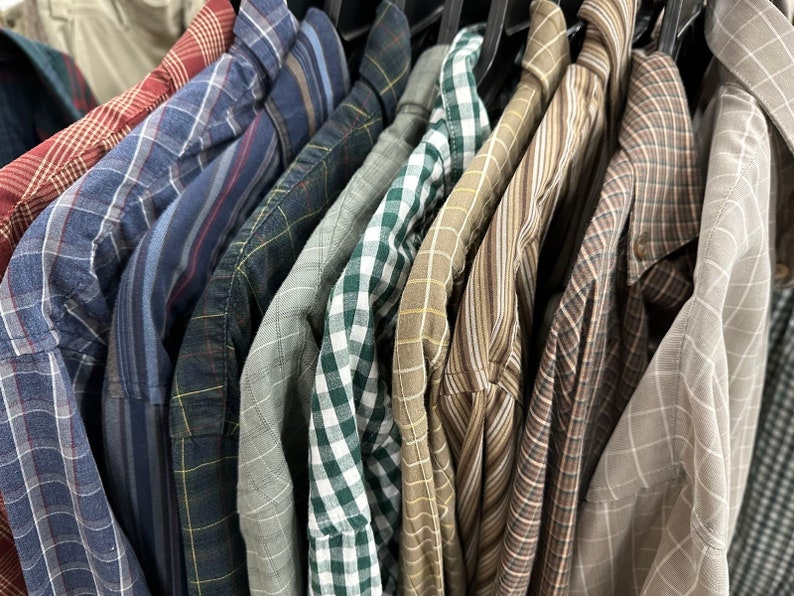 Op de afbeelding: Een verzameling geruite overhemden in verschillende kleuren en patronen, hangend aan hangers. De overhemden zijn allemaal met lange mouwen en hebben een knoopsluiting.