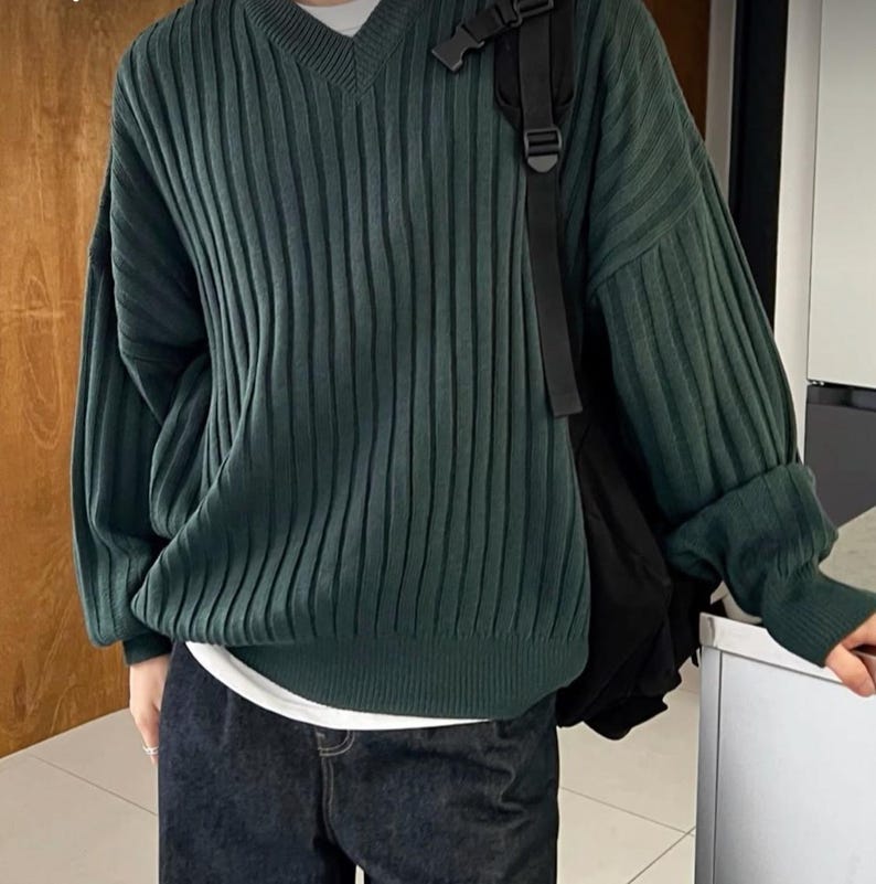 May include: A dark green, ribbed knit sweater with a V-neck. The long-sleeved sweater has a relaxed fit and is paired with dark wash denim jeans. A black shoulder bag is visible.