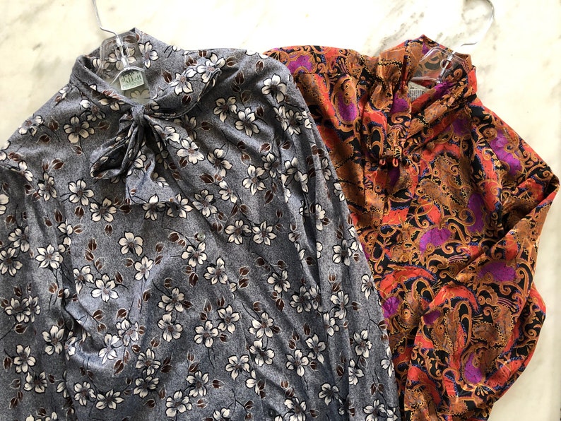 May include: Two long-sleeved blouses, one with a floral print in shades of gray and white, and the other with a paisley print in shades of orange, purple, and black.