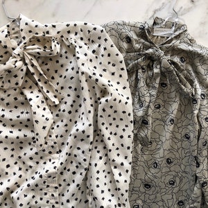 May include: Two long-sleeved blouses with tie necklines. The blouse on the left has a white background with black polka dots. The blouse on the right has a green background with a black floral pattern.