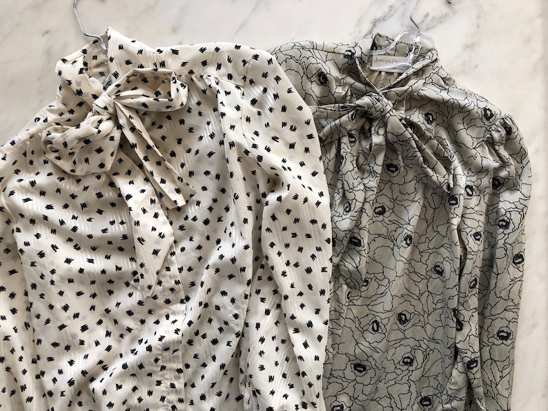 May include: Two long-sleeved blouses with tie necklines. The blouse on the left has a white background with black polka dots. The blouse on the right has a green background with a black floral pattern.