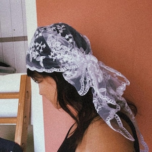 May include: A white lace head covering with floral patterns. The head covering is draped over the head and tied at the back. The background is a solid orange-red color.