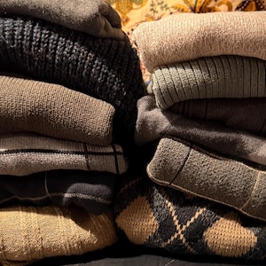 May include: Two stacks of folded sweaters in various colors and patterns. The left stack features a dark navy blue knit sweater, a tan sweater, a gray plaid sweater, and a tan argyle sweater. The right stack includes a beige sweater, a green ribbed sweater, and a gray sweater.