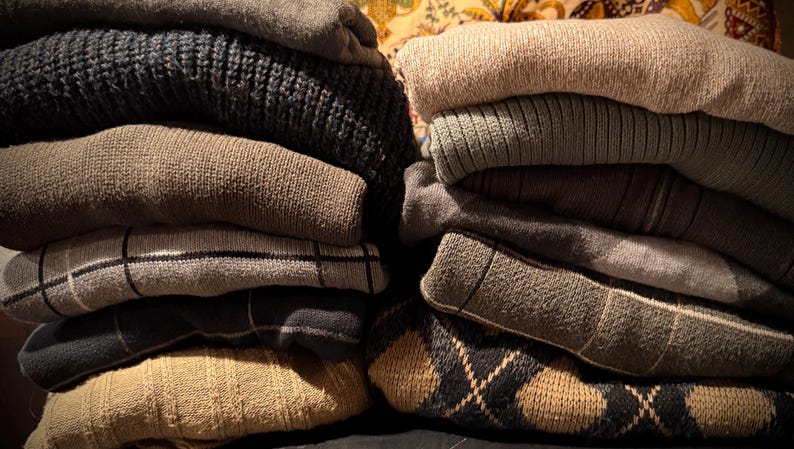 May include: Two stacks of folded sweaters in various colors and patterns. The left stack features a dark navy blue knit sweater, a tan sweater, a gray plaid sweater, and a tan argyle sweater. The right stack includes a beige sweater, a green ribbed sweater, and a gray sweater.