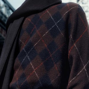 May include: A brown argyle sweater with navy blue and red accents. The sweater is paired with a black belt and dark denim jeans. A dark scarf is draped over the shoulders.