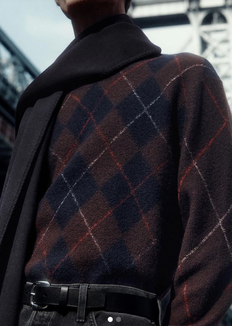 May include: A brown argyle sweater with navy blue and red accents. The sweater is paired with a black belt and dark denim jeans. A dark scarf is draped over the shoulders.