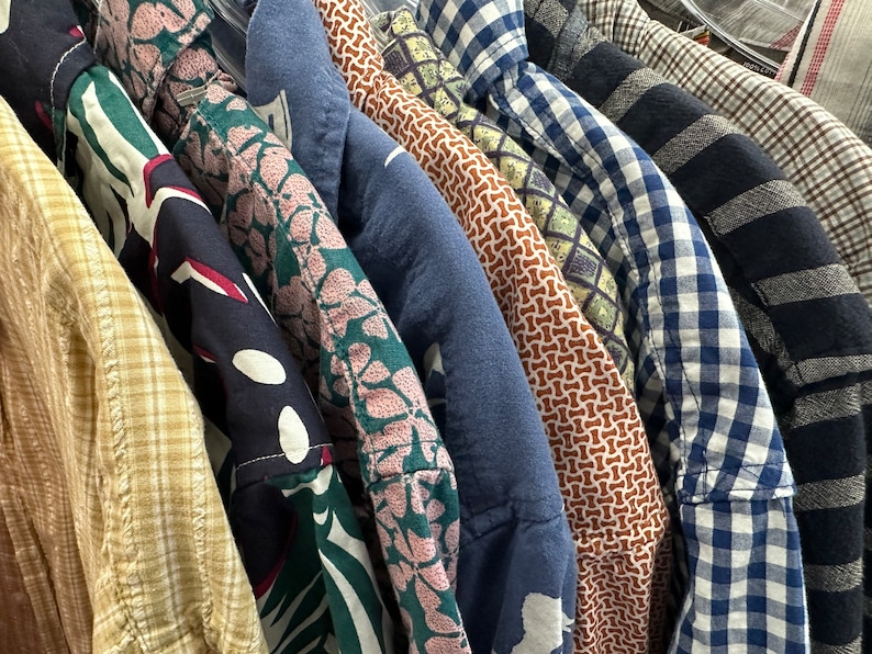 Op de afbeelding: Een verzameling van gepatroneerde overhemden in verschillende kleuren, waaronder blauw, groen, bruin en wit. De overhemden hangen aan een rek en zijn in een rij gerangschikt.
