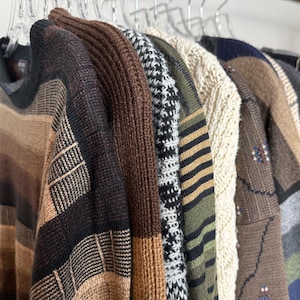 May include: A collection of sweaters hanging on clear hangers. The sweaters display a variety of patterns, including stripes, houndstooth, and cable knit. Colors range from brown and beige to green and black, showcasing a range of textures and styles.