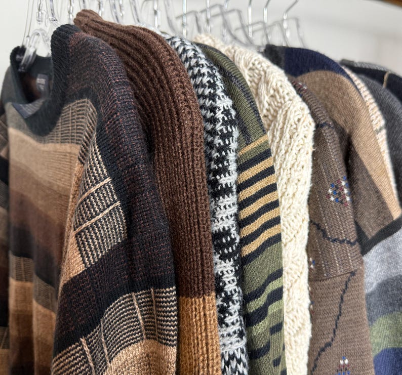 May include: A collection of sweaters hanging on clear hangers. The sweaters display a variety of patterns, including stripes, houndstooth, and cable knit. Colors range from brown and beige to green and black, showcasing a range of textures and styles.