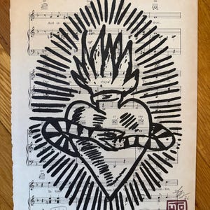May include: Black and white Sacred Heart art print on vintage sheet music. The artwork features a stylized heart with flames and rays, and the sheet music has musical notes and text. The print is signed and dated.