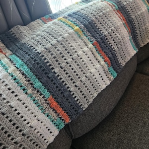 May include: A handmade crochet blanket in shades of gray, white, and blue, with stripes of orange and teal. The blanket is draped over a gray sofa, showcasing its textured stitch pattern and color variations. A cozy, decorative home textile.
