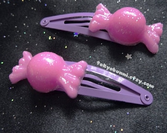 May include: Two purple plastic hair clips with pink candy-shaped charms. The charms have a glittery finish.