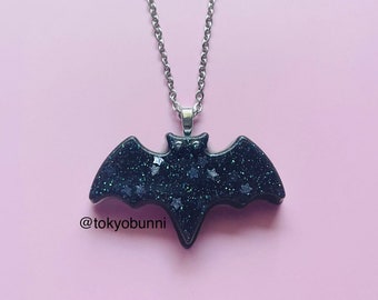 May include: A black bat pendant necklace with a silver chain. The bat-shaped pendant is dark with glitter and star accents. The text "@tokyobunni" is below the pendant against a pink background.