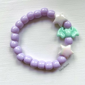 May include: A beaded bracelet featuring light purple barrel beads. The bracelet includes a light green bat charm and two star charms in white and pink. The text "Tokyobunni" is visible on the bracelet.