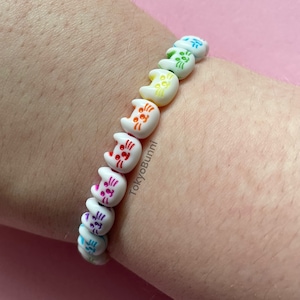 Könnte beinhalten: Ein farbenfrohes Armband mit weißen, katzenförmigen Perlen mit bemalten Details in verschiedenen Farben. Die Perlen sind aufgefädelt und bilden ein verspieltes Accessoire. Das Armband wird an einem Handgelenk gezeigt.
