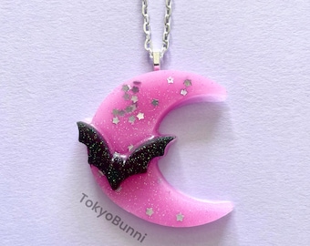 May include: A silver chain necklace with a pink crescent moon pendant. The moon is decorated with silver glitter and a black bat. The pendant is about 2 inches in diameter.