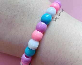 May include: A colorful beaded bracelet featuring a variety of pastel-colored beads, including pink, purple, blue, and white. The bracelet is worn on a wrist and has the text "Tokyobunni" printed on it.