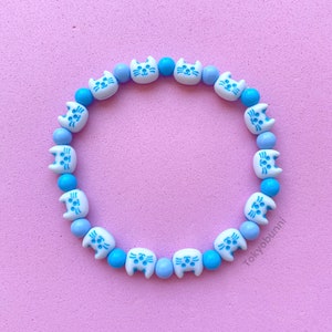 May include: A white and blue beaded bracelet featuring cat faces. The beads alternate between blue and white, with the cat faces on the white beads. The bracelet is displayed against a pink background.