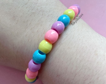 Op de afbeelding: Een kleurrijke kralenarmband met roze, gele, blauwe en paarse kralen. De armband is geregen en de tekst "Tokyobunni" is zichtbaar op de armband.