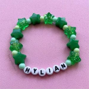 Può includere: Un braccialetto di perline a forma di stella verde con il nome "HYLIAN" scritto in perline bianche.