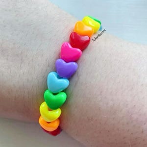 Puede incluir: Un brazalete con cuentas en forma de corazón de colores del arcoíris. Las cuentas están dispuestas en una secuencia de rojo, naranja, amarillo, verde, azul, morado y rosa. El texto "Tokyobunni" es visible en la imagen.