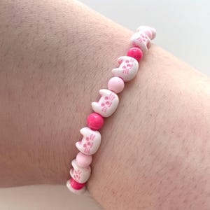 May include: A pink and white beaded bracelet with cat face charms. The bracelet is made with small, round beads and features a repeating pattern of pink and white beads.