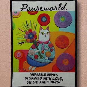 May include: A colorful fabric art piece featuring a whimsical cat holding a bouquet. The artwork includes the text "Pauseworld" and the quote "WEARABLE WHIMSY. DESIGNED WITH LOVE, STITCHED WITH 'OOPS.'" The cat is seated on a sofa with colorful circular designs.