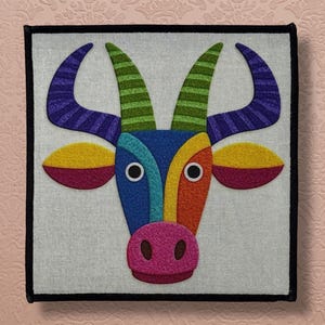 Può includere: Un'opera d'arte tessile quadrata e colorata raffigurante una testa di toro stilizzata. Il toro ha sezioni blu, arancioni, gialle e viola, con corna a strisce verdi e viola e un naso rosa. L'opera è incorniciata in nero.