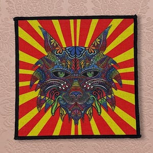 May include: A square patch featuring a detailed, colorful illustration of a lynx's face. The lynx is depicted with intricate patterns in shades of blue, green, orange, and purple, set against a red and yellow sunburst background, framed by a black border.