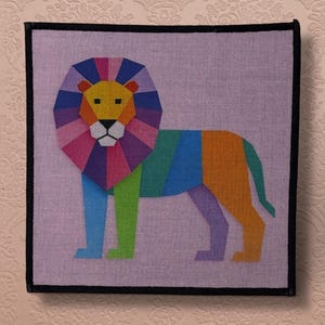 May include: A colorful geometric lion design on a square canvas. The lion features a rainbow mane and body, with a black border. The background is a light purple color.